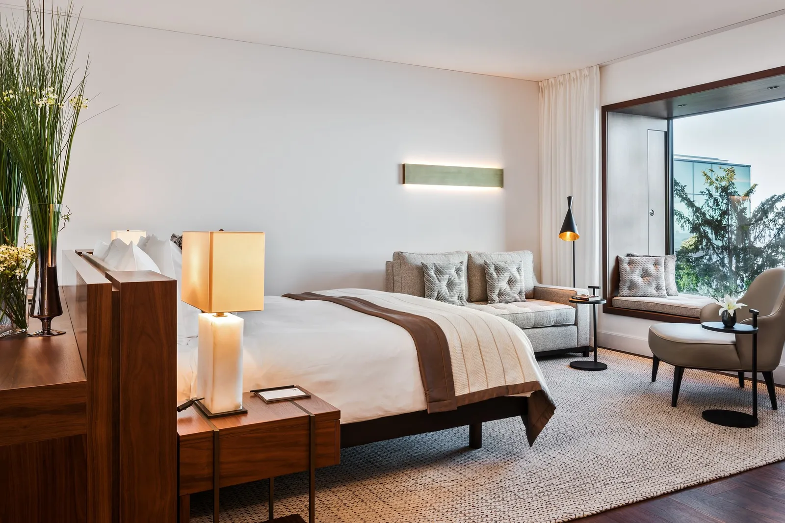 The bedroom of a Contemporary Lake View Panorama Suite: a bed facing the room, a sofa and an armchair by a large window onto the trees