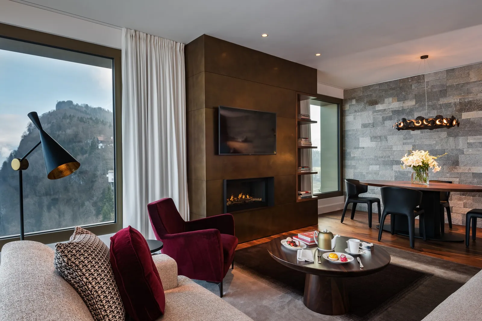The living room of a Contemporary Lake View Premium Suite: a red armchair, a fireplace in a bronze wall, a dining table and the cliff in the window