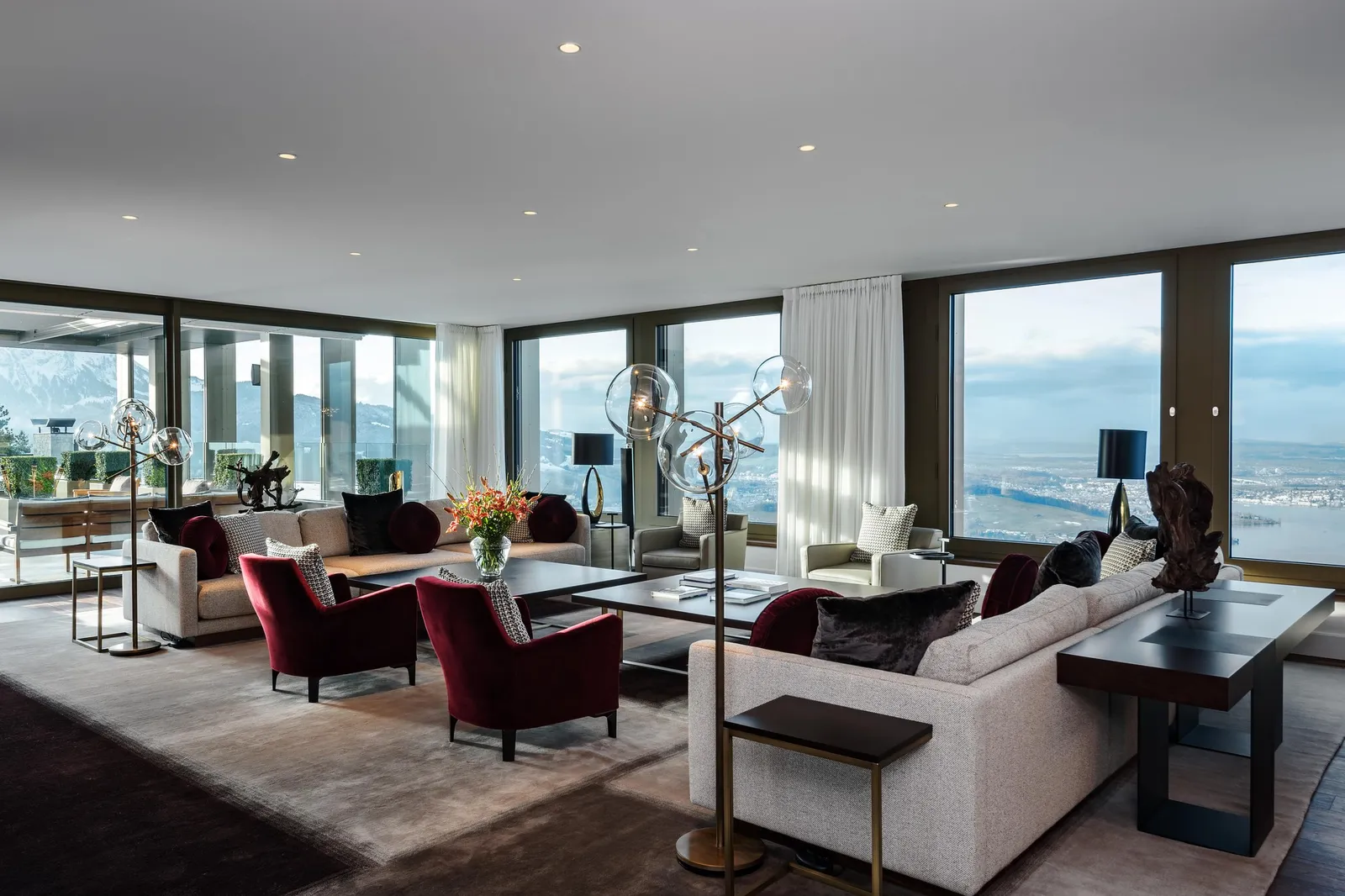 The Royal Suite’s living room: sofas and red armchairs round low tables, glass lamps, and the lake through windows on 3 sides