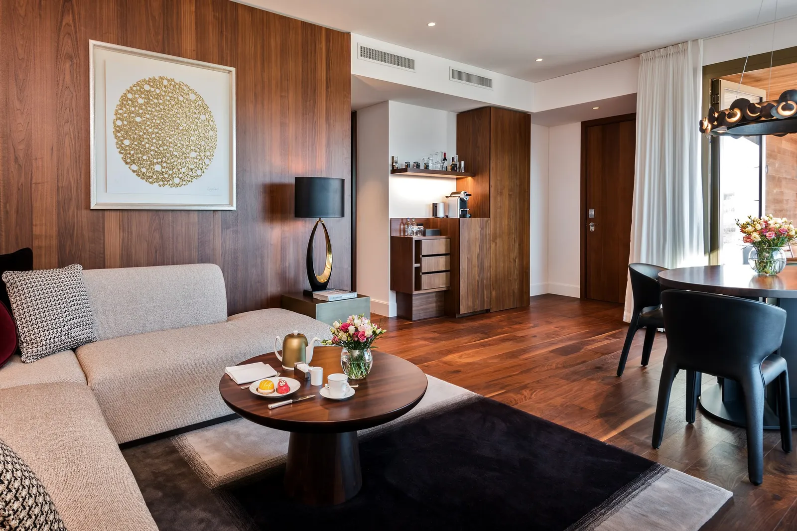 The living room of a Contemporary Lake View Spa Suite: walnut walls, a gold artwork, a sofa and a round table, and a dining table by the window