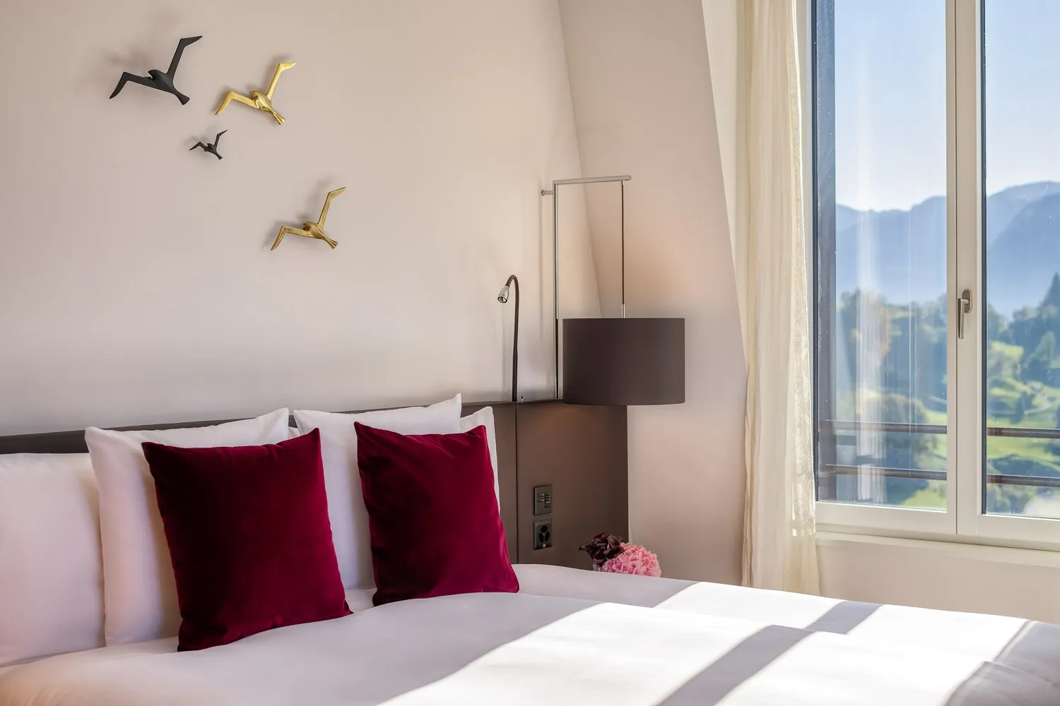 A Heritage Alpine Deluxe Room: a bed with red velvet cushions under metal birds on the wall, and green hills in the tall window