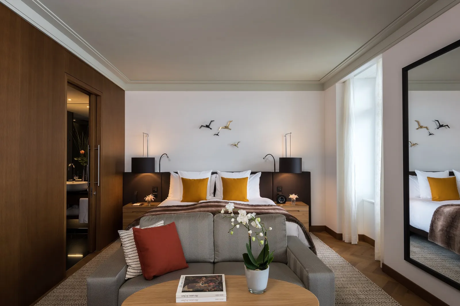 A Heritage Lake View Deluxe Room: a bed with mustard cushions, a grey sofa with a red cushion, and birds in flight on the wall