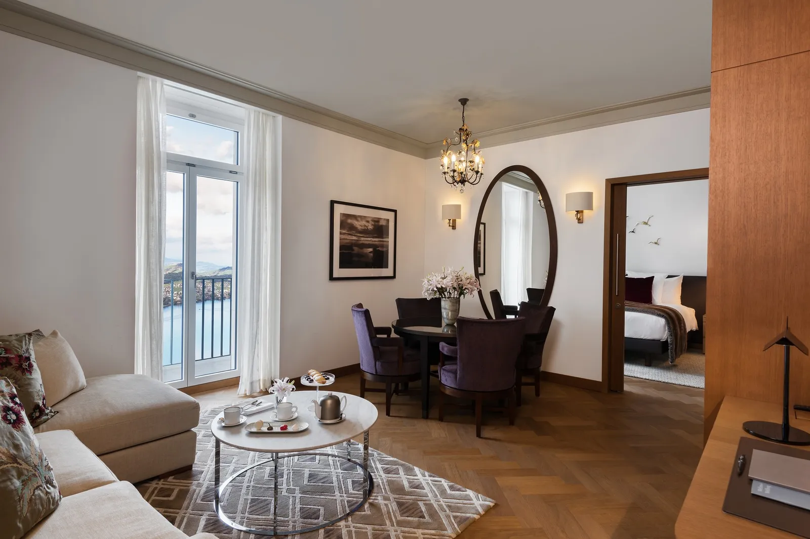 The living room of a Heritage Lake View Suite: an oval mirror, a small chandelier, a dining table and French windows onto the lake