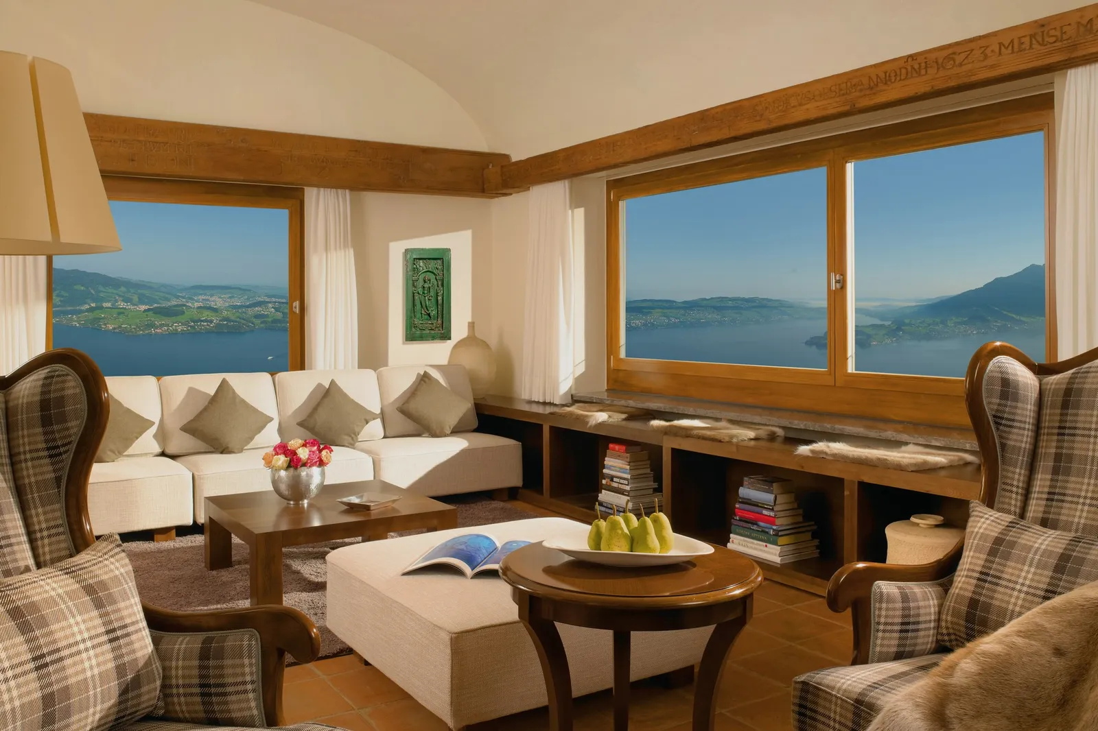 The Lake View Cottage’s living room: tartan wing chairs, a white sofa, a vaulted ceiling with a carved beam, and the lake in 2 wide windows