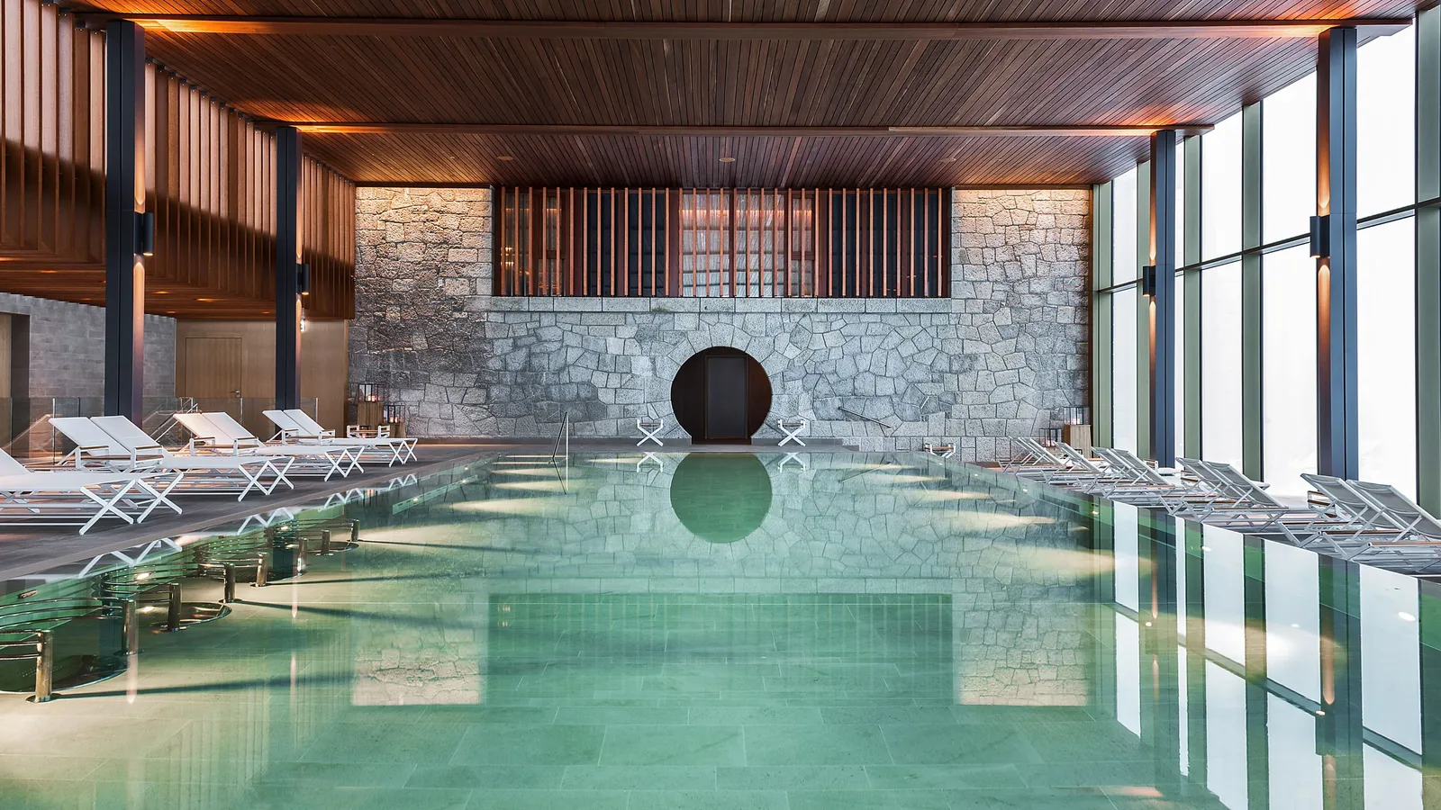 The Alpine Spa’s indoor pool: green water, a wall of rough stone with a round dark door, white loungers and a timbered ceiling