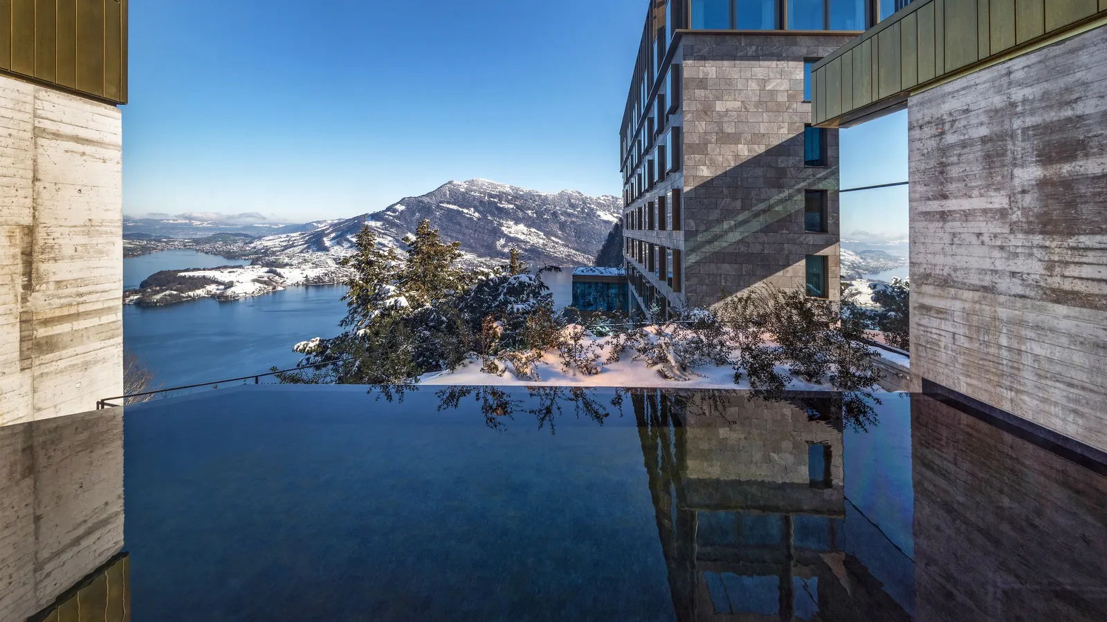 The infinity pool in winter between the concrete walls of the spa, with snow on the trees and the lake and the white mountains beyond the edge