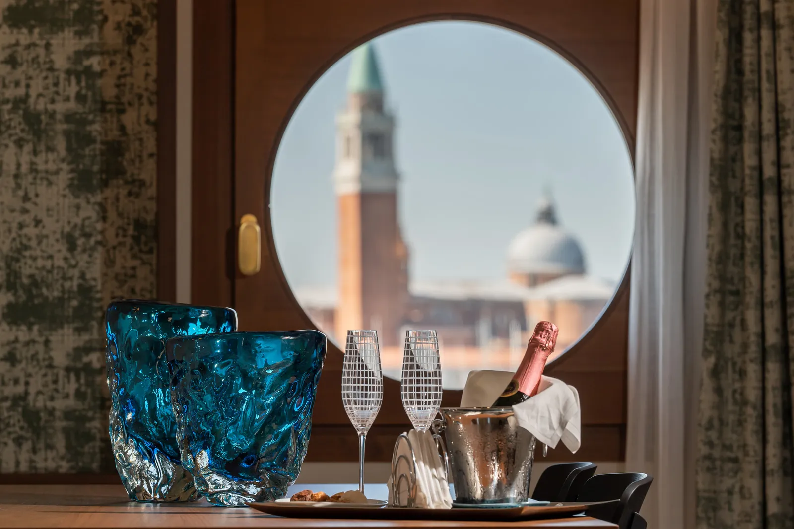 A round window frames the bell tower and dome of San Giorgio Maggiore; on the sill, 2 blue glass vases and 2 flutes