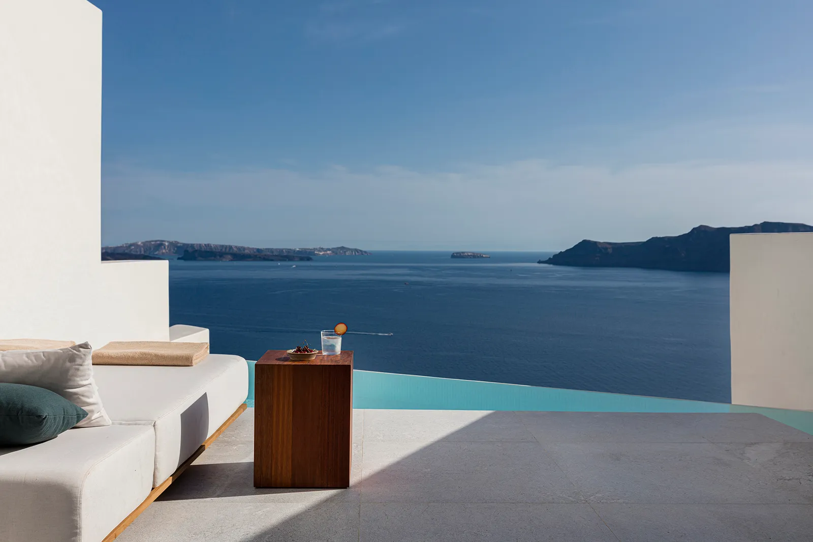 A daybed with cushions, a wooden side table and the edge of an infinity pool facing the caldera.