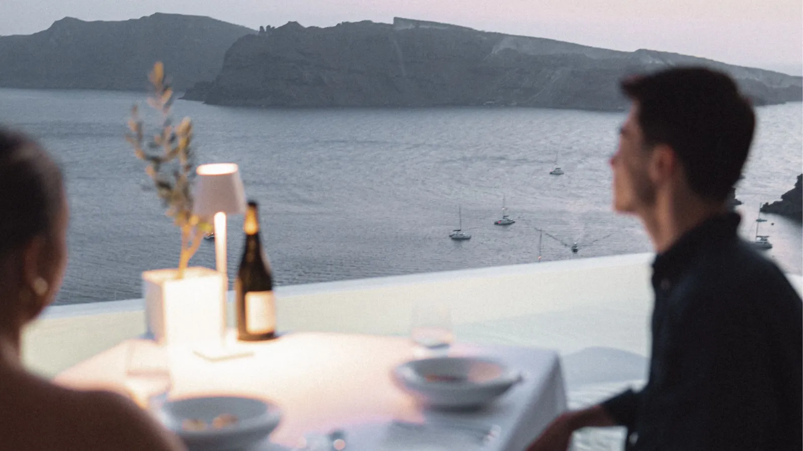 Dinner for 2 at dusk: the table with a lamp and a bottle of wine, and boats on the caldera beyond.