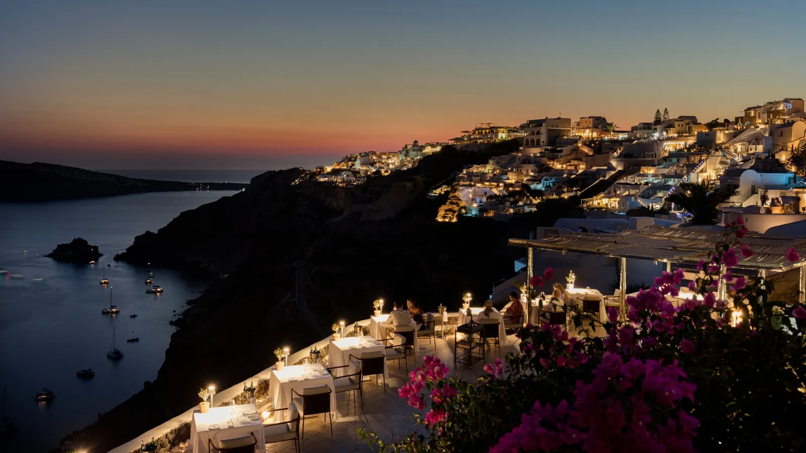 Petra’s tables laid on the terraces at dusk, bougainvillea in the foreground, the lights of Oia on the cliff and boats in the bay.