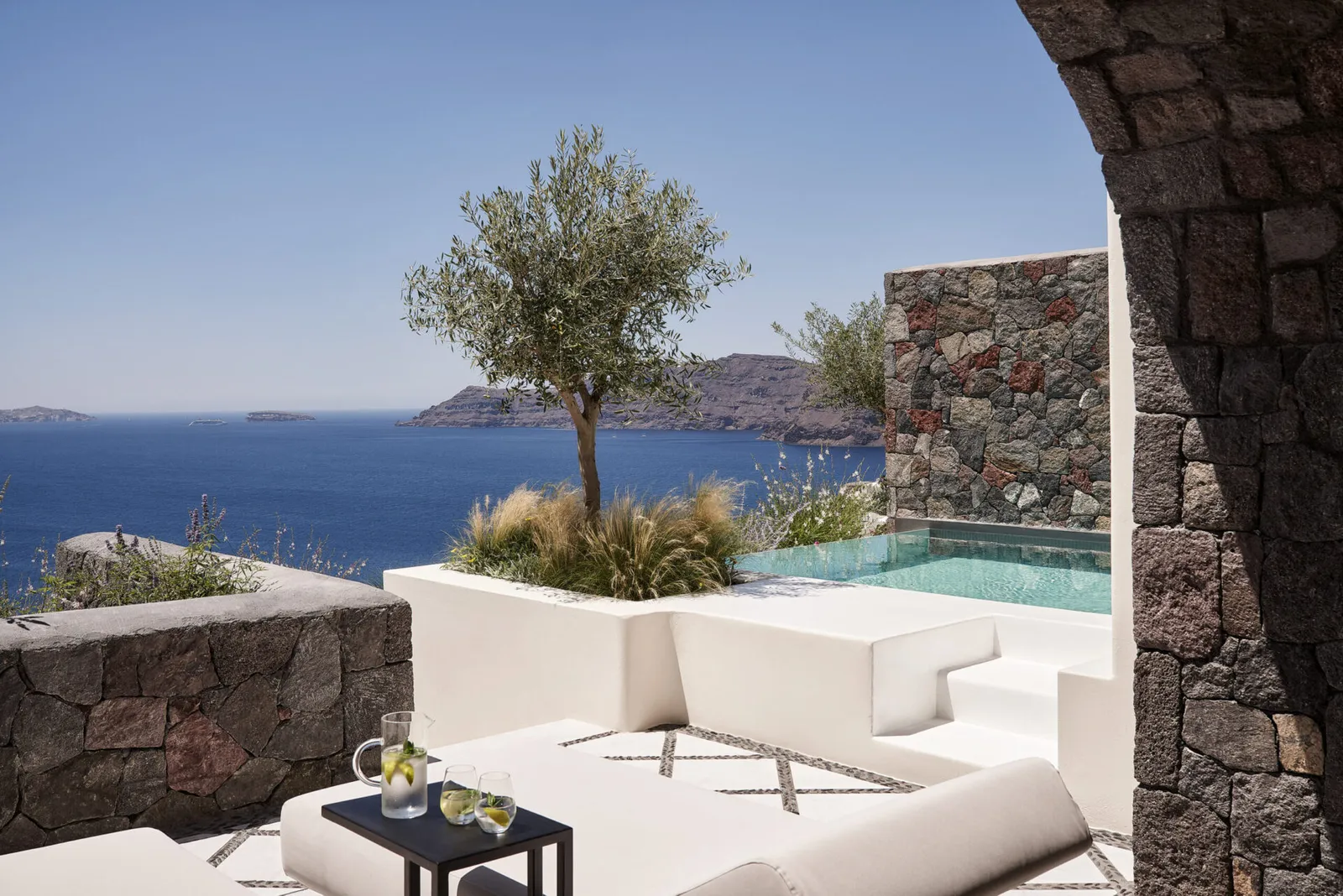A terrace with an olive tree, lava-stone walls, white sofas and a pool, seen through an arch.