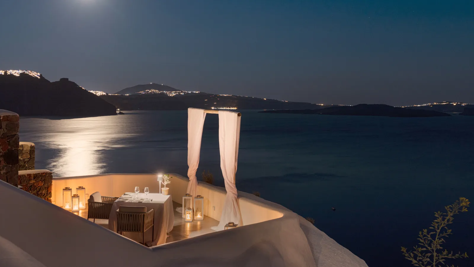 The Panorama Balcony at night: a single table laid between lanterns and 2 white drapes, with the moon on the sea and the lights of the island.