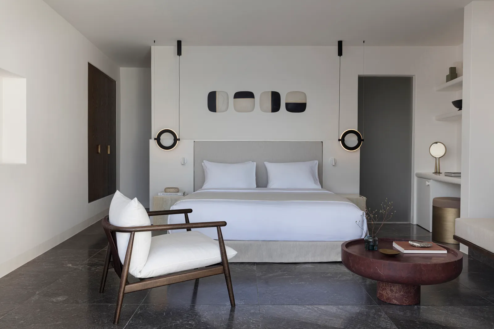 A bedroom in grey tones: the white bed, a wooden armchair, a dark red table and pendant lamps.