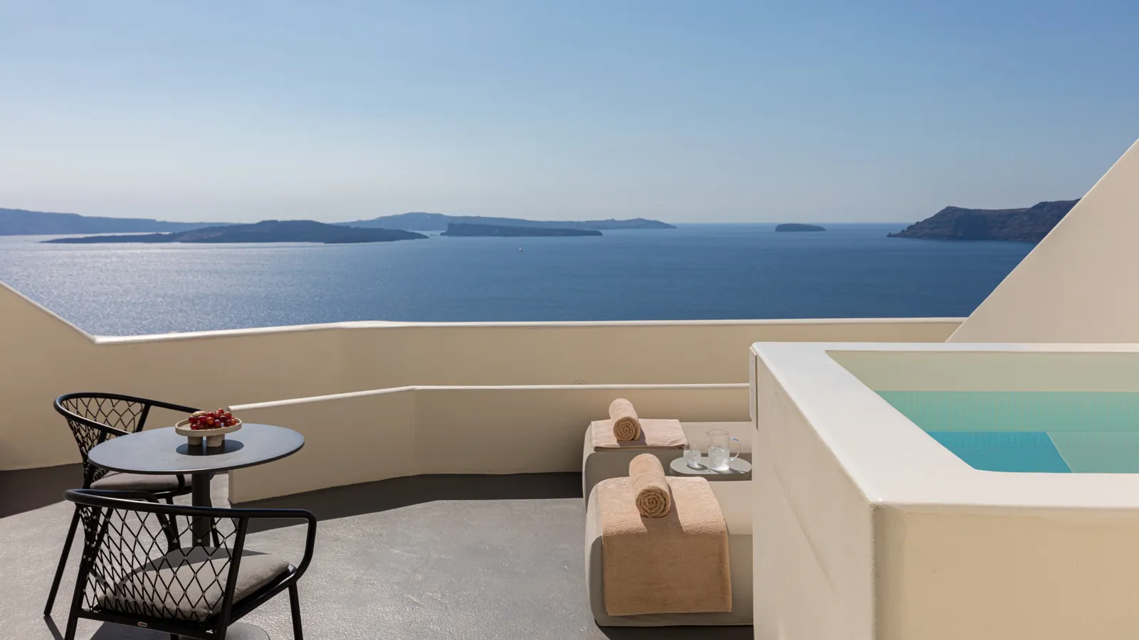 A Junior Suite terrace: a table with 2 black chairs, 2 loungers and the plunge pool, above the sea of the caldera.