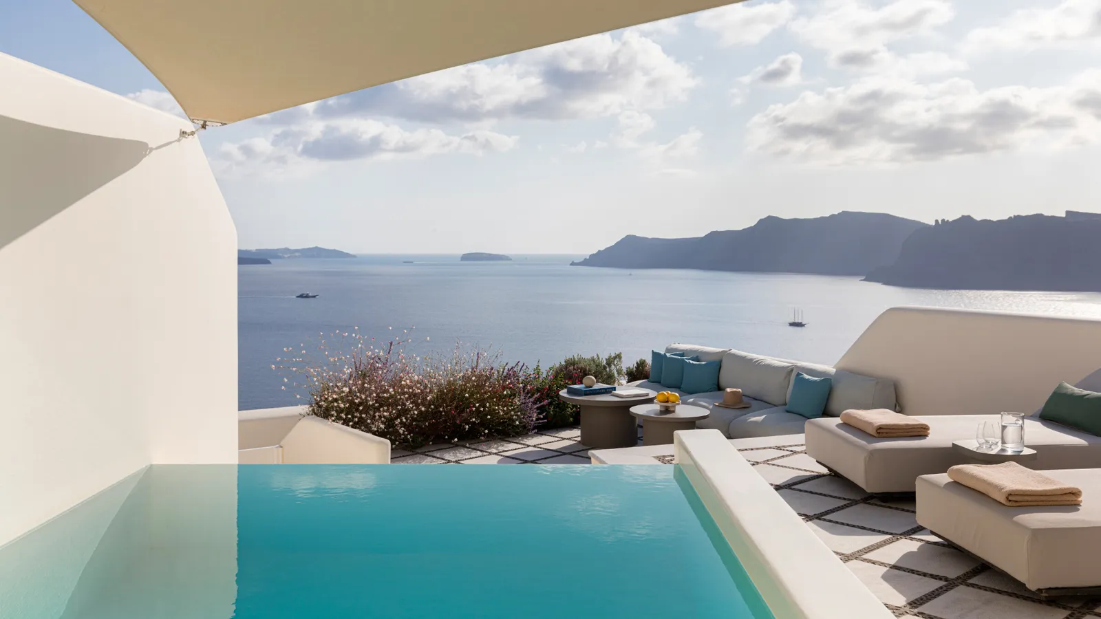 The Royal Pool Suite’s terrace: the pool, loungers with blue cushions under a sail and clouds above the caldera.