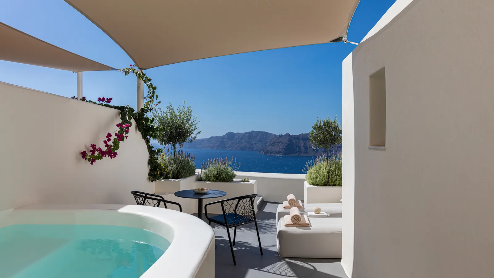 A Superior Suite terrace: the curved plunge pool under a shade sail, bougainvillea, a table and the caldera.