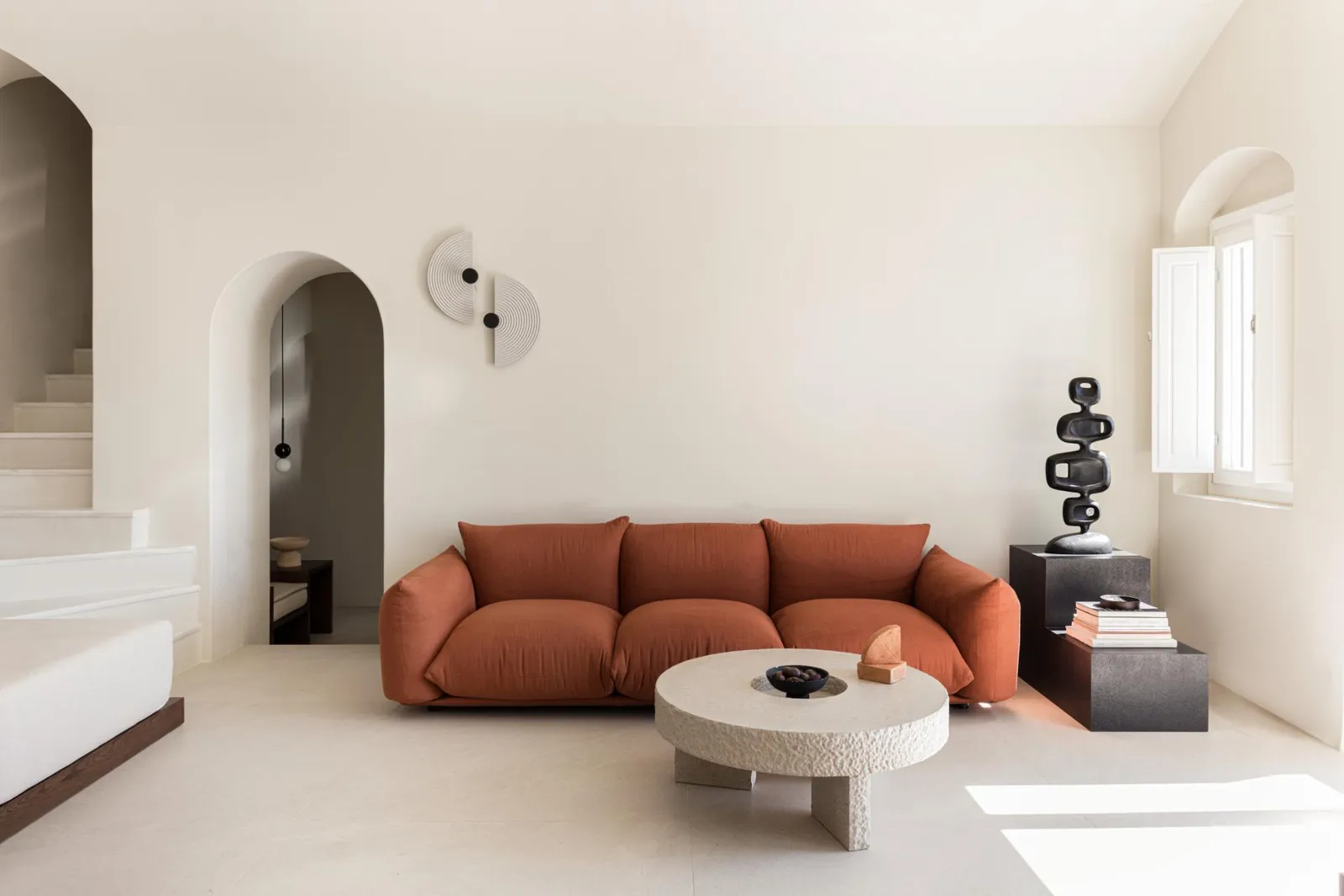 The Three Bedroom Villa’s living room: a rust-coloured sofa, a round stone table and white walls with arched niches.