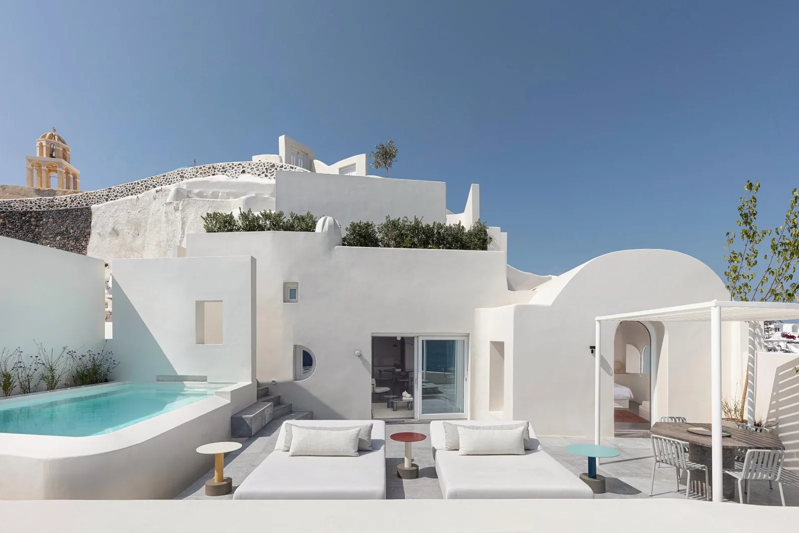 The Two Bedroom Pool Villa from outside: white volumes, the terrace with its sofas, the pool and an ochre church on the left.