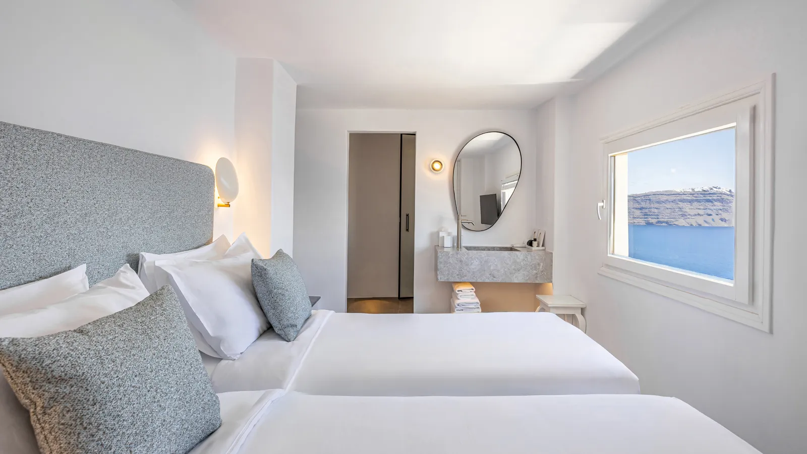 A bedroom of the Two Bedroom Suite: the bed with its grey-blue headboard, a dressing table with an oval mirror and a window onto the houses of Oia.