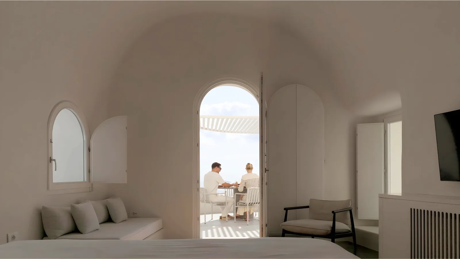 The vaulted interior of a suite, with a white sofa and the arched door open onto the terrace, where 2 guests are having breakfast.
