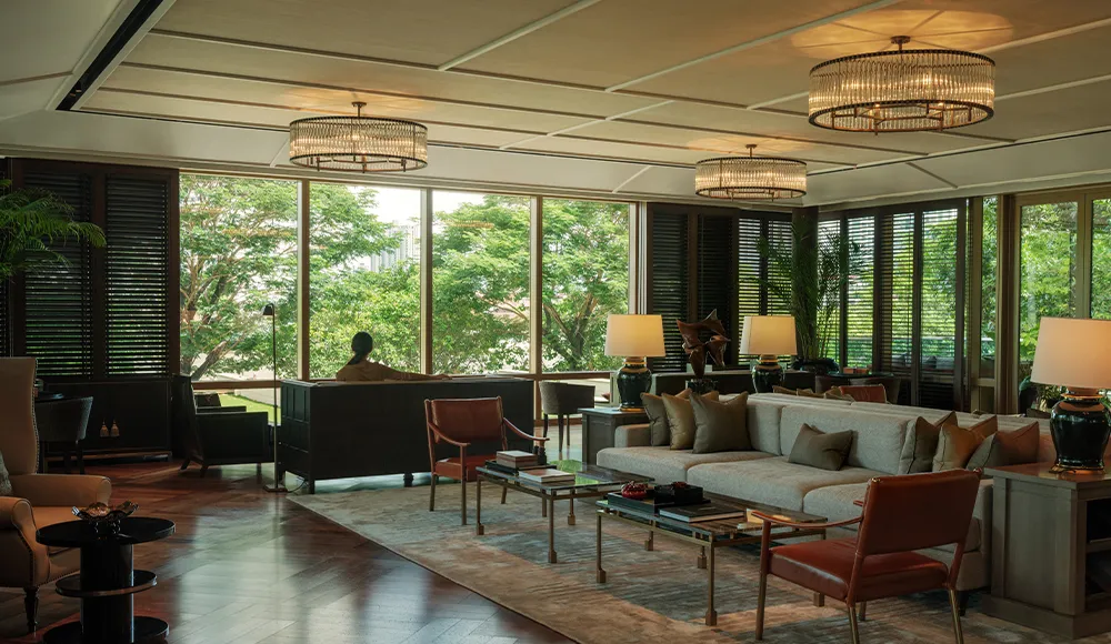 La Living Room: divani e poltrone su un tappeto, lampadari bassi e una parete di finestre sugli alberi del lungofiume.