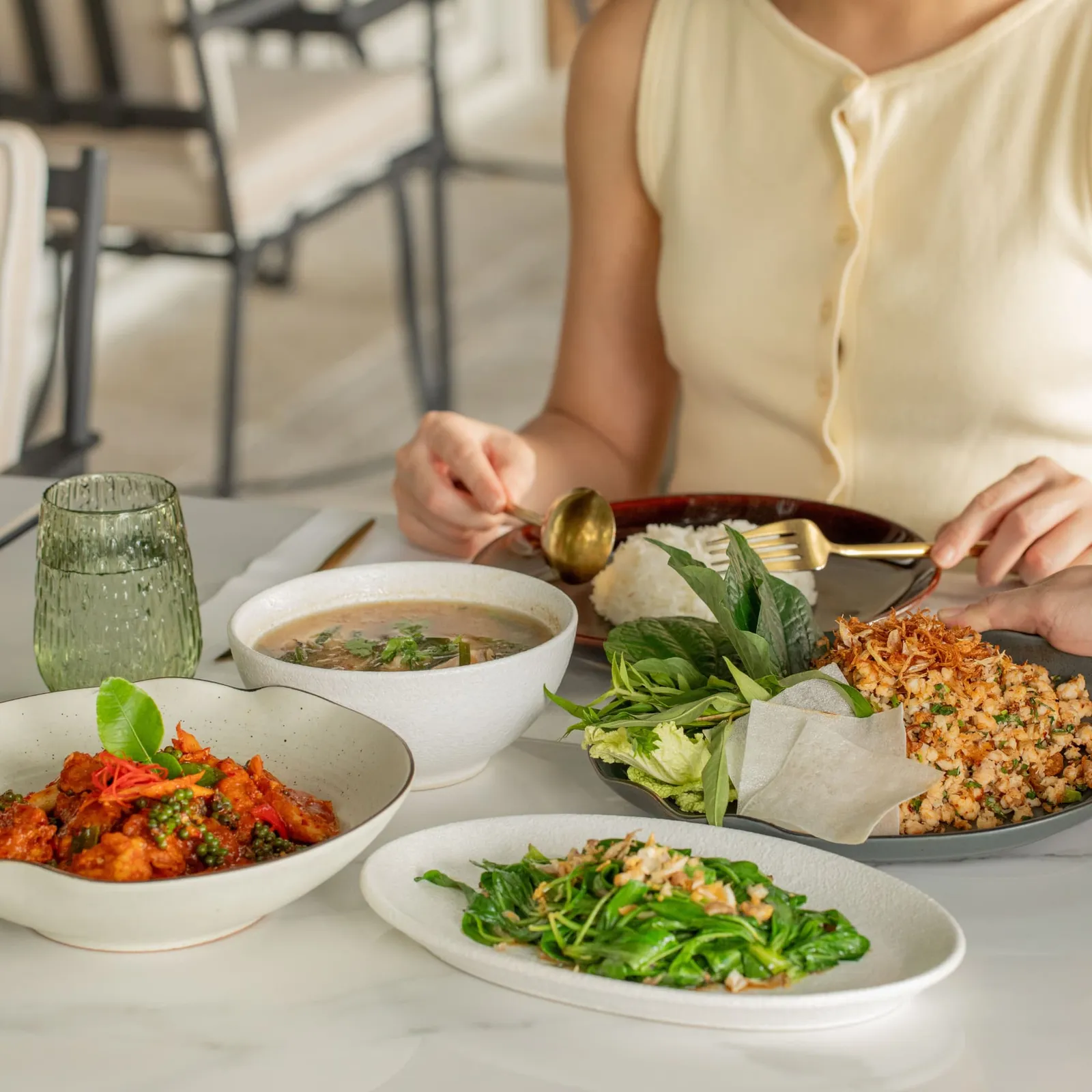 Piatti thai su un tavolo bianco: un curry rosso, verdure saltate, una zuppa e un piatto di erbe fresche, una mano che serve.