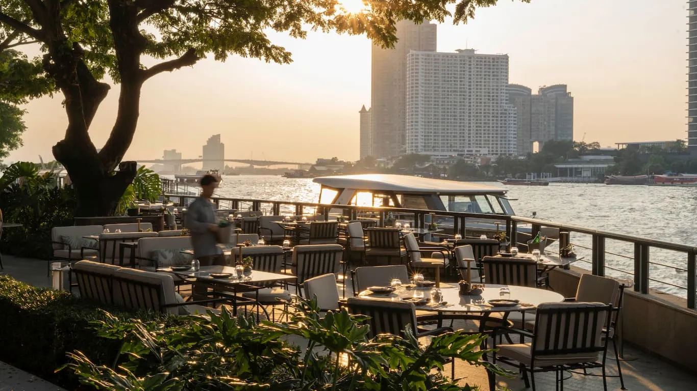 La terrazza sul Chao Phraya al tramonto: tavoli apparecchiati sotto un grande albero, la barca dell’hotel ormeggiata alla ringhiera e i grattacieli sull’altra riva.