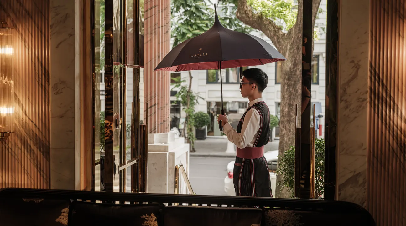 A doorman in livery holding a red umbrella at the hotel’s glass door, the street behind him