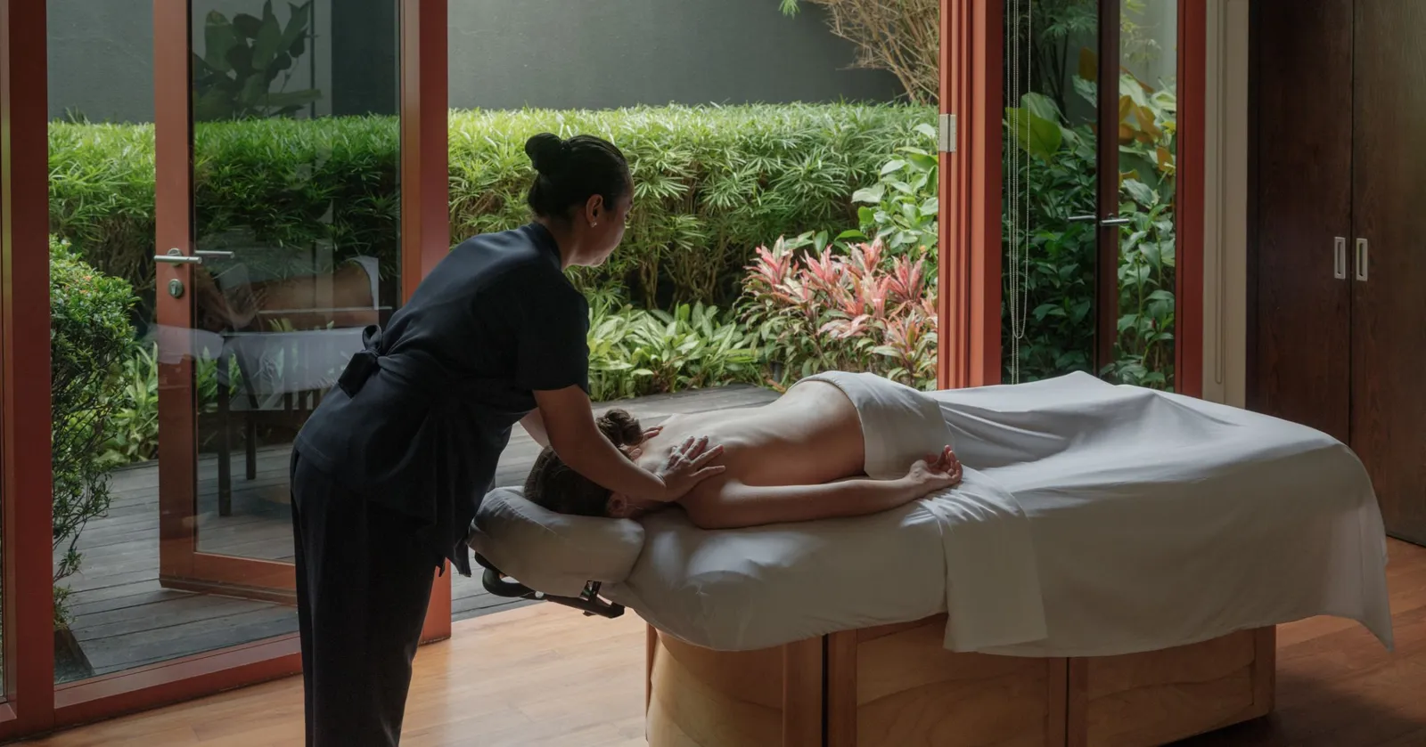 A treatment room at Auriga Spa: a therapist at work on a guest lying on the table, the doors open on a private garden.