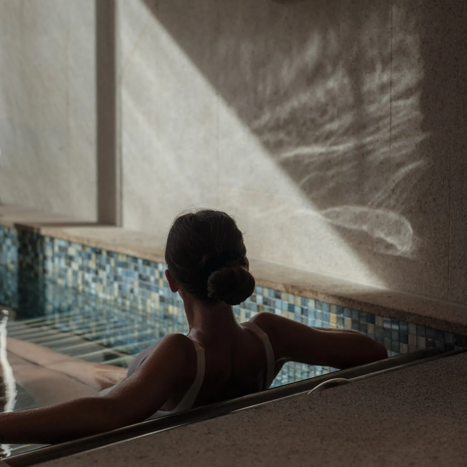 A guest from behind in Auriga Spa’s vitality pool, mosaic at the waterline and light falling across the wall.