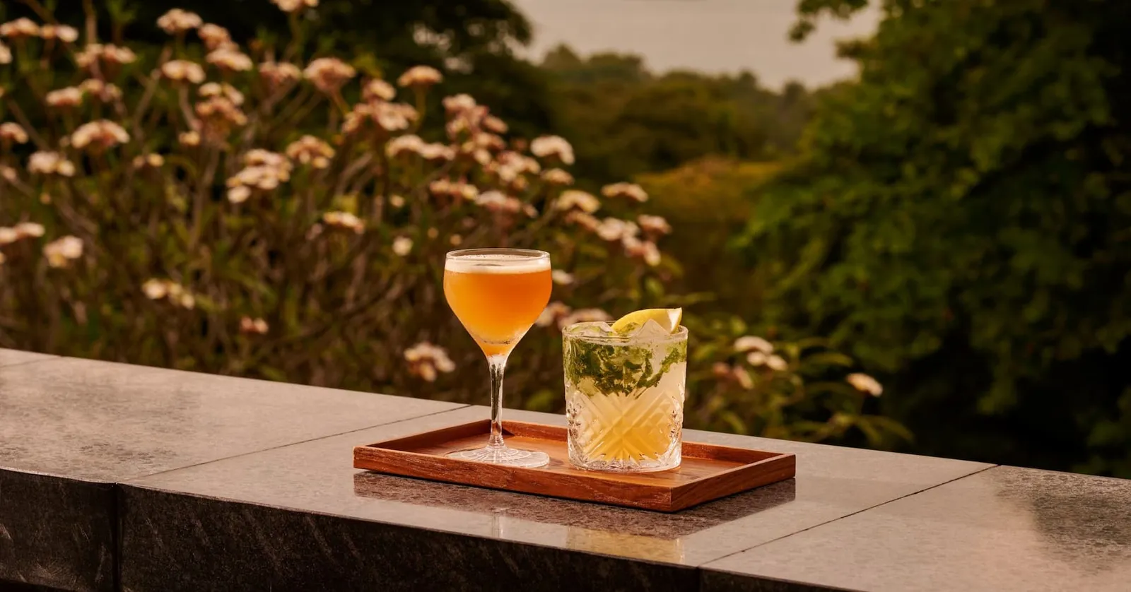 2 cocktails on a wooden tray on the terrace ledge of Bob’s Bar, flowering shrubs and trees behind in evening light.