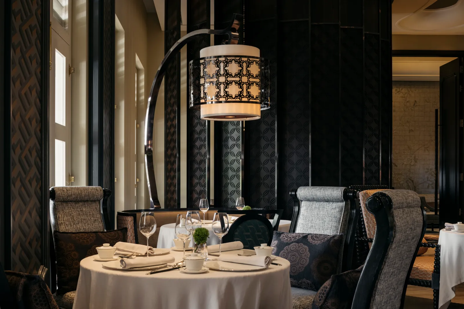 Cassia’s dining room: a round table laid with white linen, high-backed chairs, a patterned lantern above and black lacquered columns.