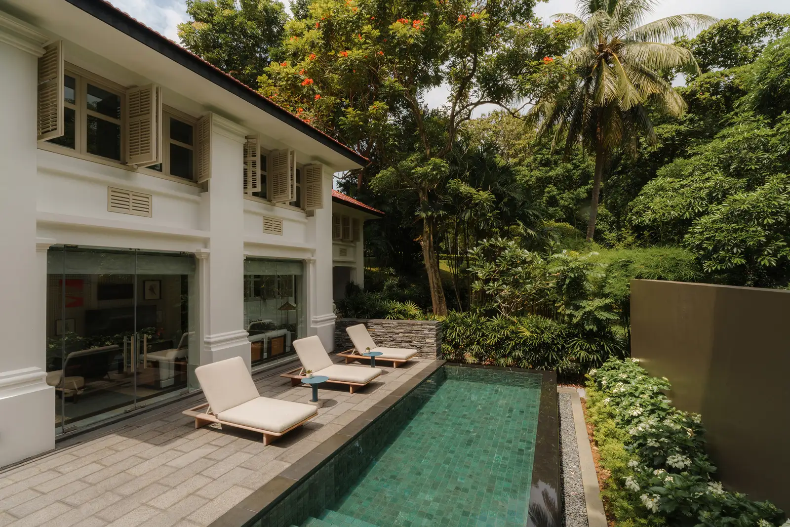 A Colonial Manor’s pool: a long, narrow pool beside the white bungalow, 3 sunbeds on the deck and a slope of garden.