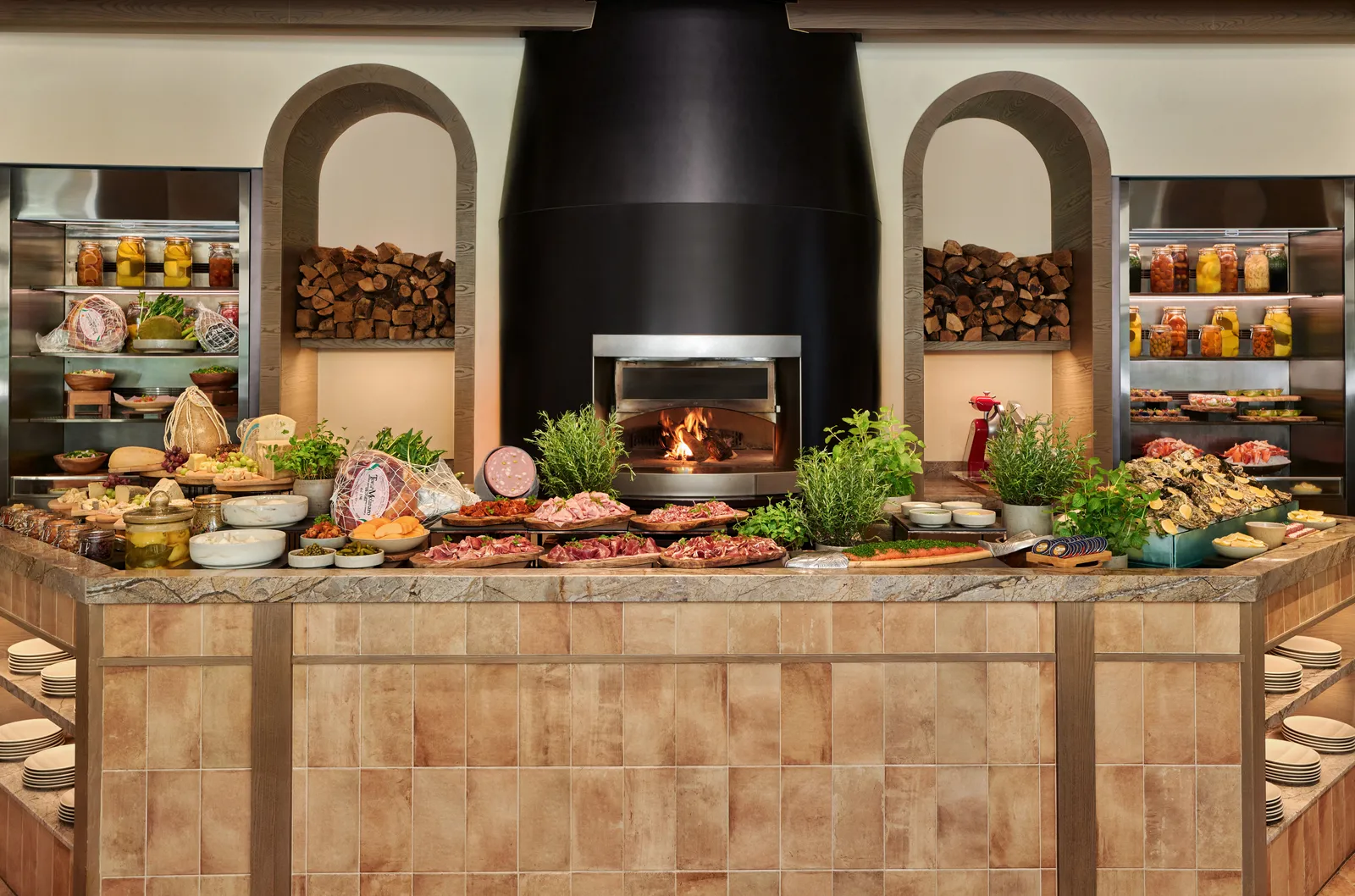 Fiamma’s counter laid for the Sunday brunch: cured meats, cheeses and jars of preserves in front of the wood fire, with logs stacked in 2 arched niches.