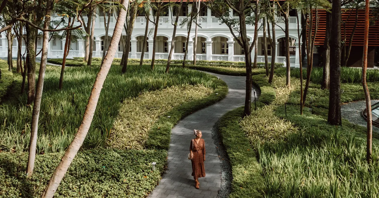 A paved path winding through grass and young trees below the colonial wing, a guest walking along it.