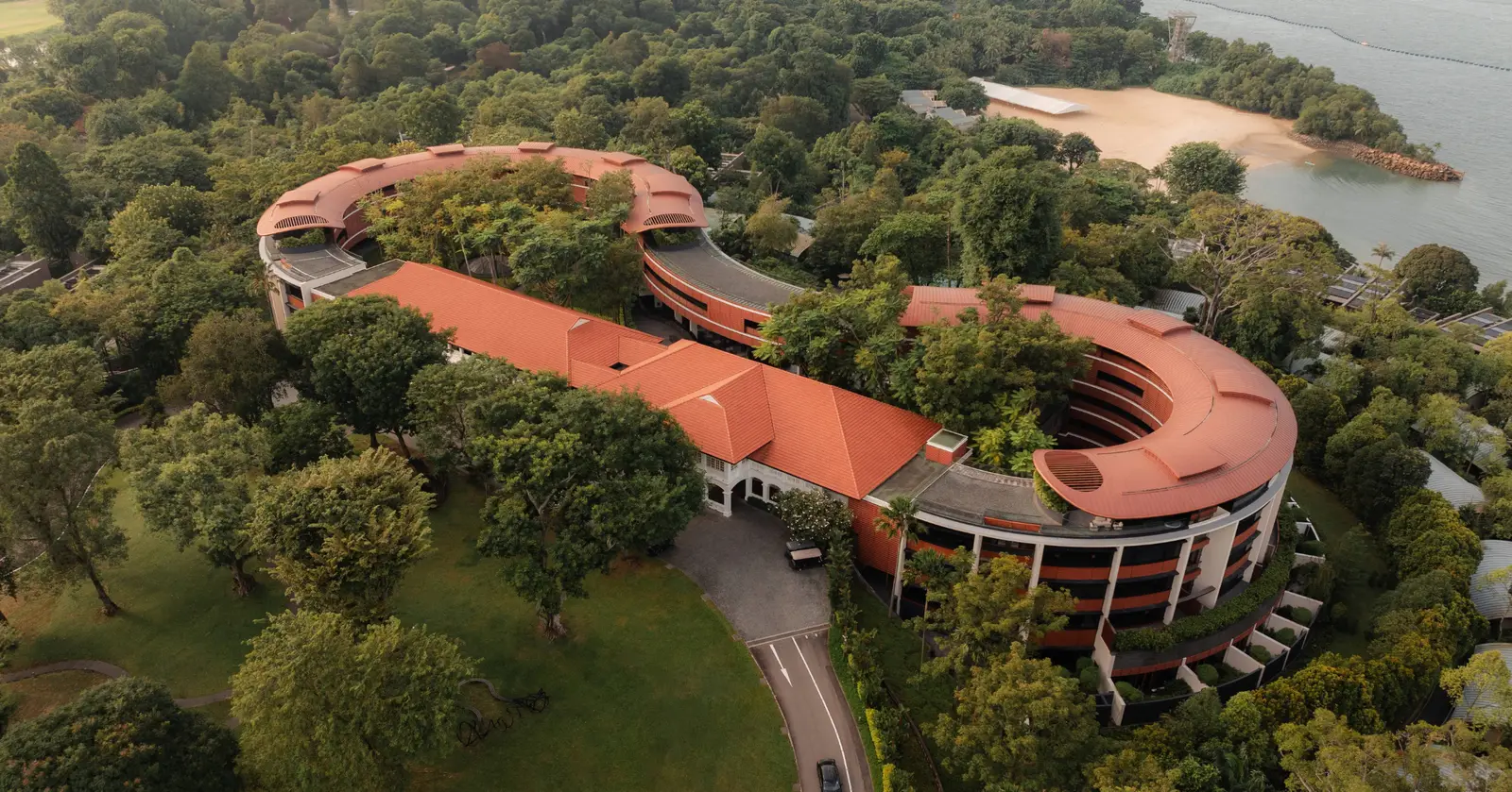 Capella Singapore from the air: the red-roofed colonial buildings joined to a contemporary wing that curves in 2 loops among the trees, with a sandy beach and the sea behind.