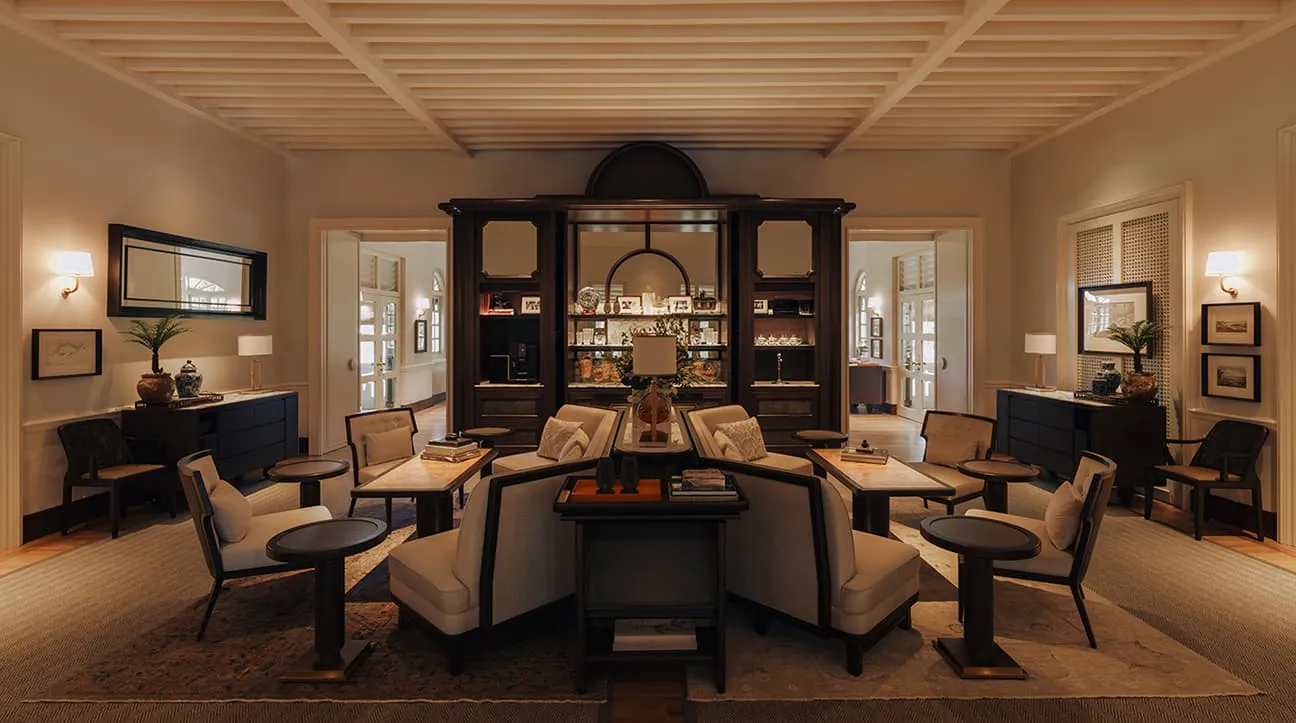 The Living Room: armchairs round low tables on a rug, a dark cabinet of books and porcelain, white shuttered doors on either side.