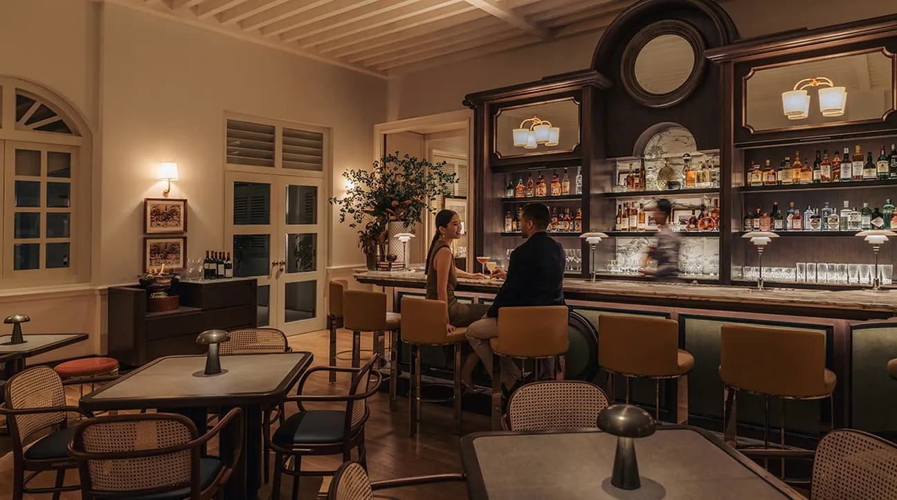 The Pineapple Room: the bar with bottles on mirrored shelves, 2 guests on stools and a bartender at work, cane chairs at the tables.