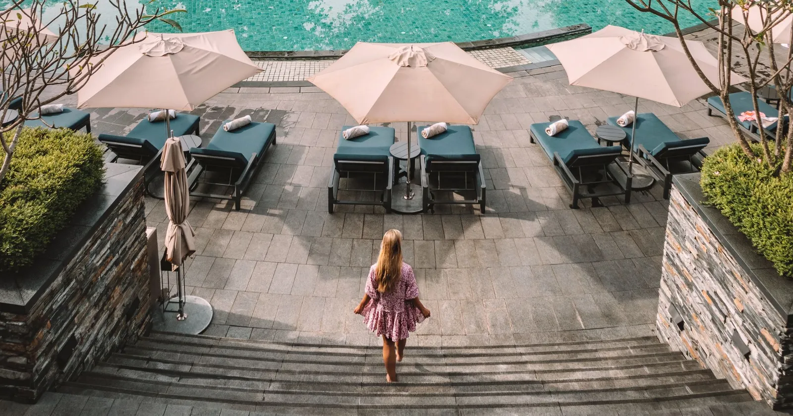 A pool terrace from above: 3 parasols over pairs of sunbeds at the water’s edge, stone steps and a guest walking down them.