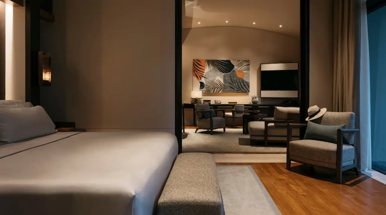 A One-Bedroom Villa: the bed in the foreground, the sitting room beyond a sliding partition and an armchair with a straw hat on it.
