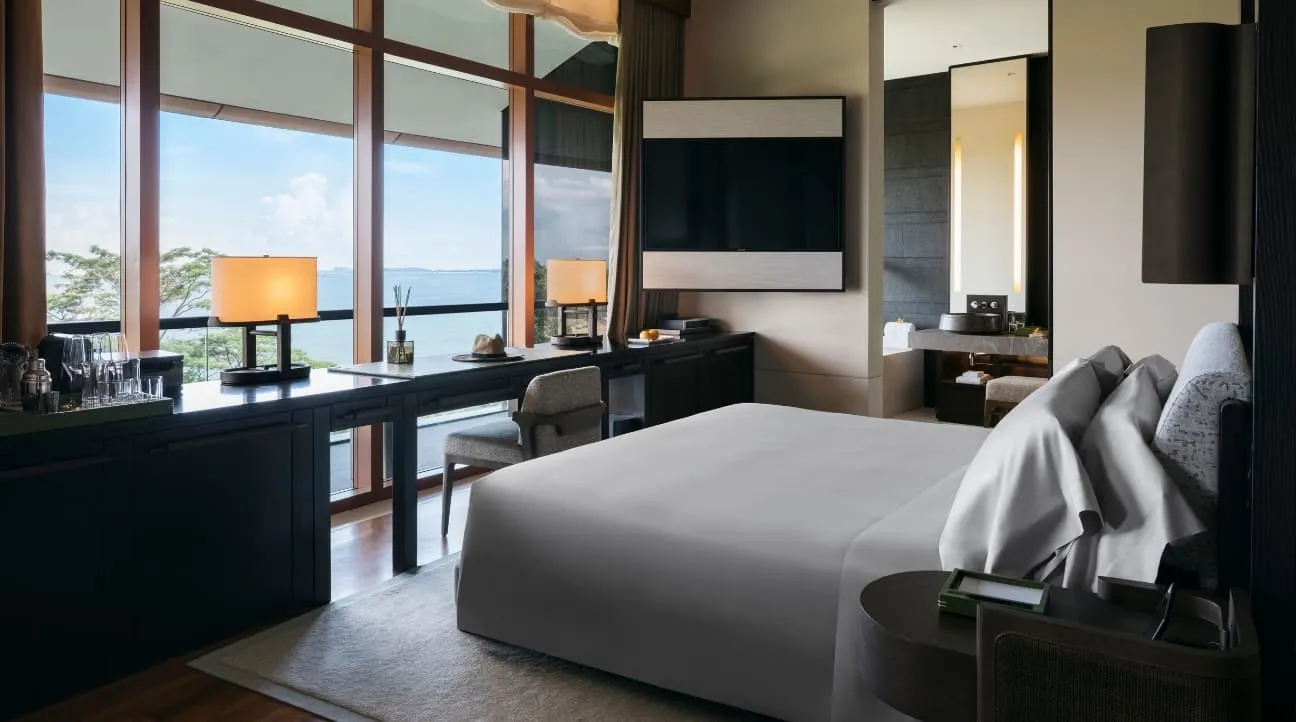 A Sentosa Suite’s bedroom: the bed facing the windows, a desk beneath them and the sea beyond the trees.