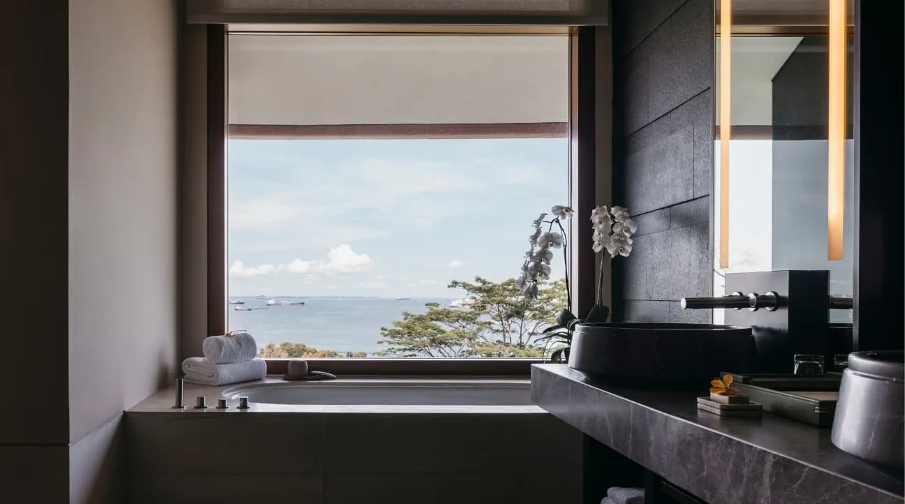 The bath of a Premier Seaview Room under a window on the treetops and the sea, a stone basin on the right.