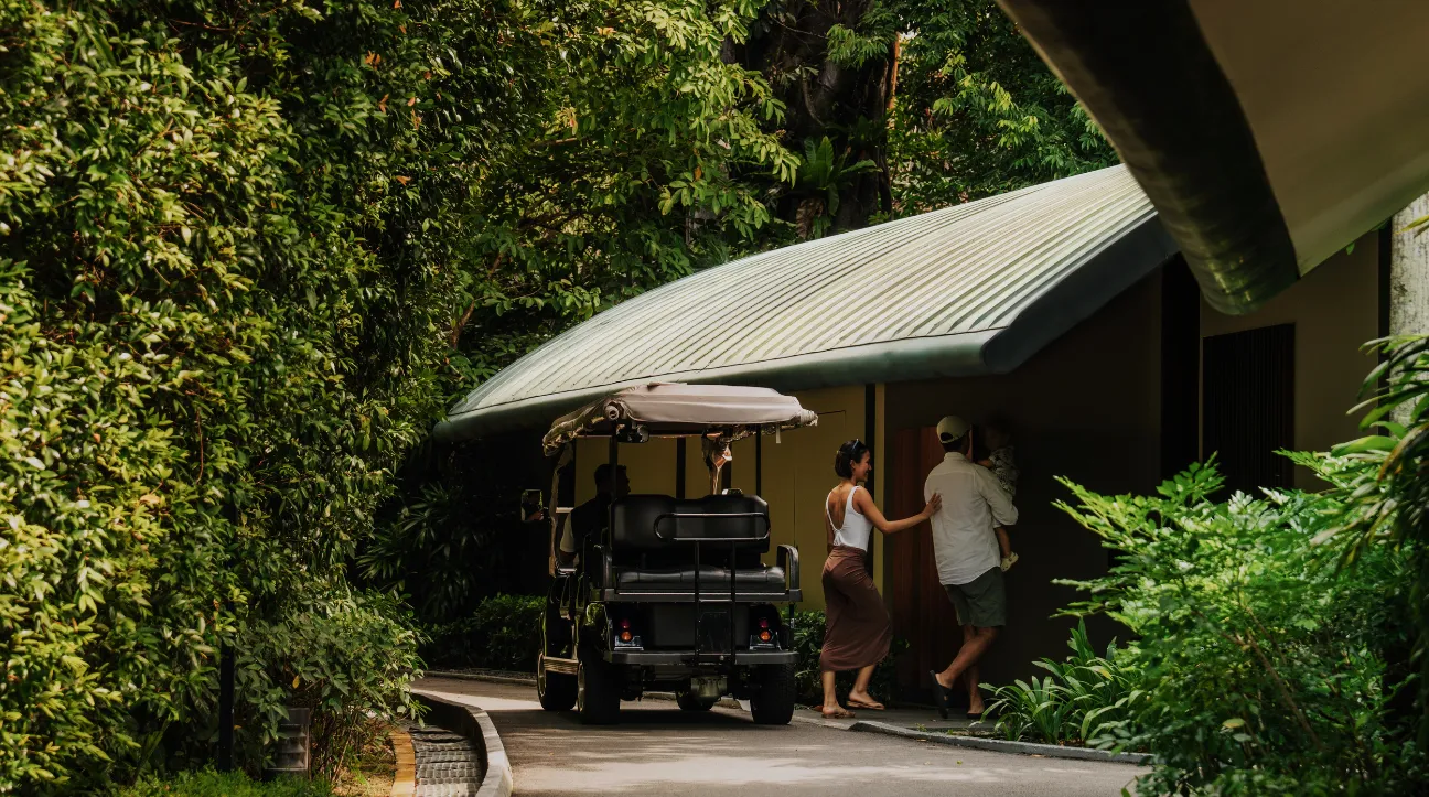 A buggy parked at a villa’s entrance among the trees, 2 guests walking to the door under the curved roof.