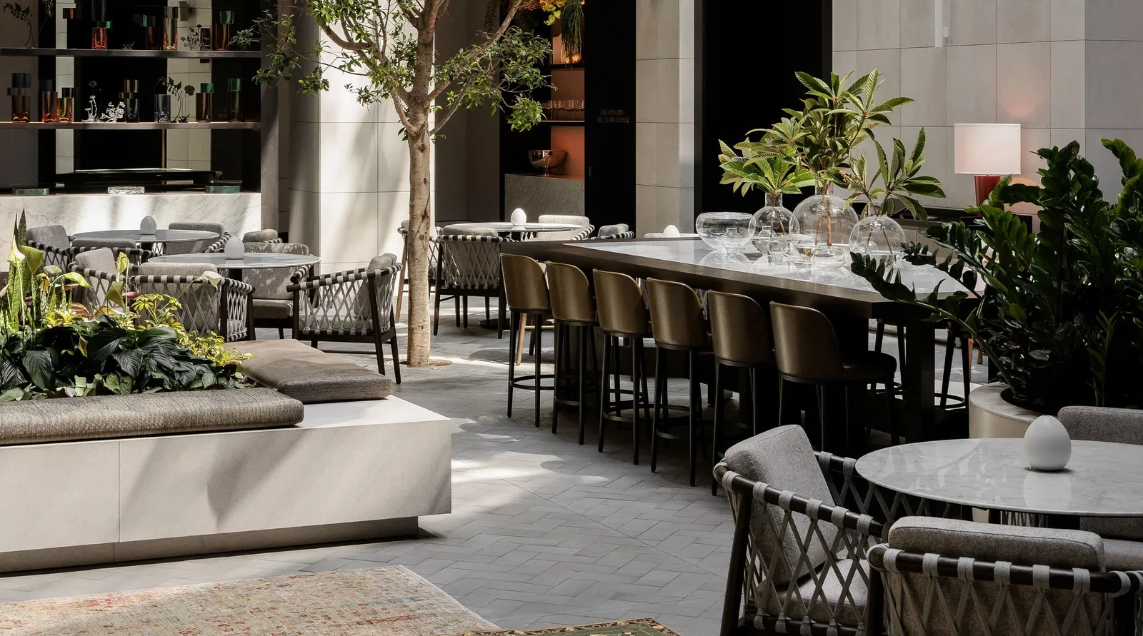 Aperture in the atrium: a long marble table with bar stools, trees and planters in daylight