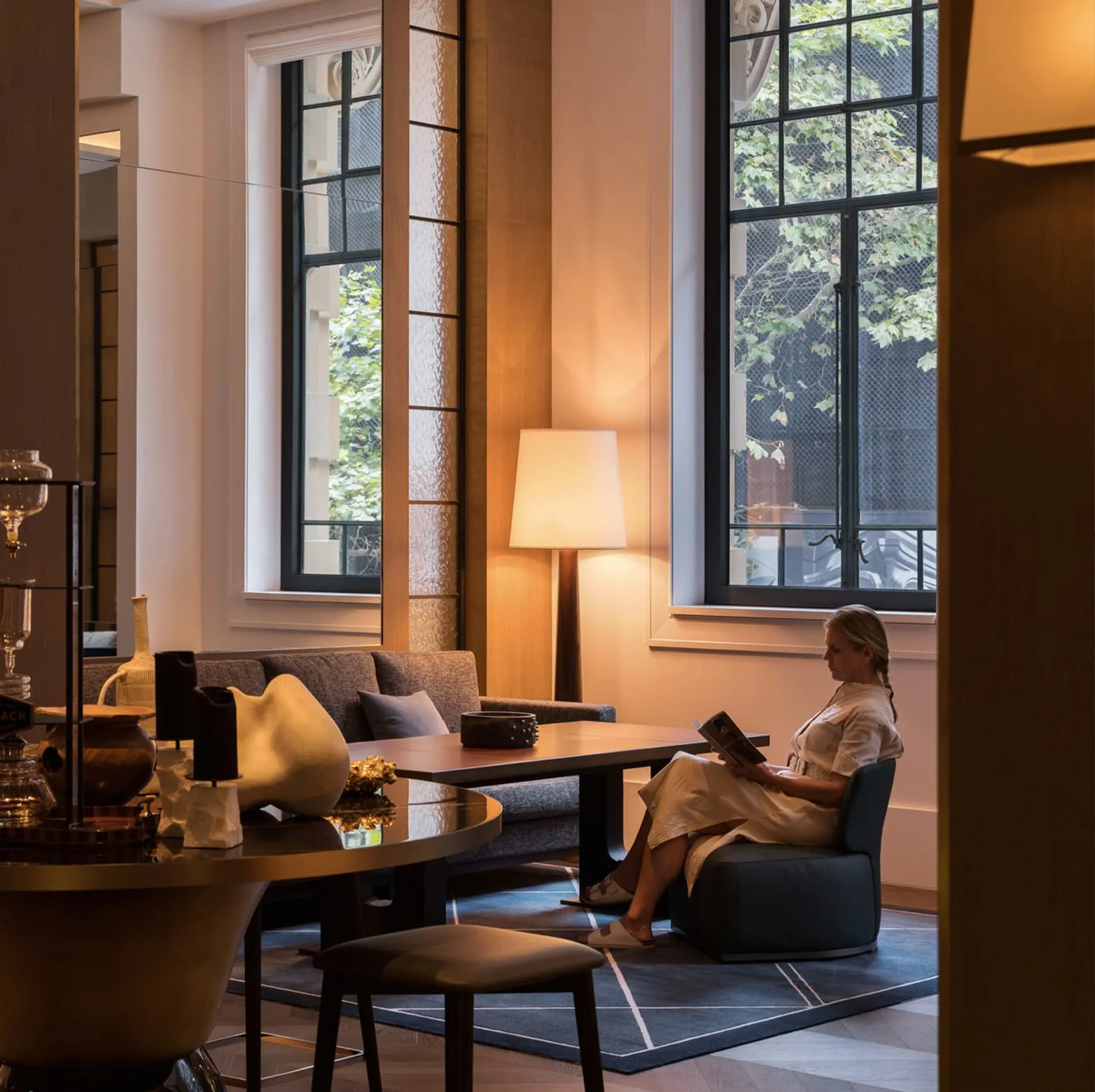 The Living Room: a guest reading in an armchair by a tall window, a lamp and a low table