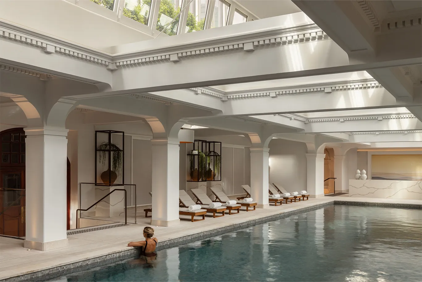 A swimmer at the edge of the indoor pool, white columns and loungers along the side, skylights above