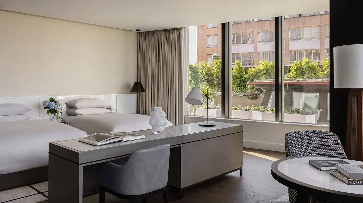 A Skyline Room: a white bed, a desk and a window onto trees and city buildings