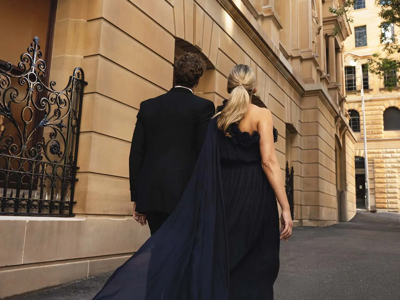 A couple in evening dress walking past the sandstone wall and wrought-iron gate of the building
