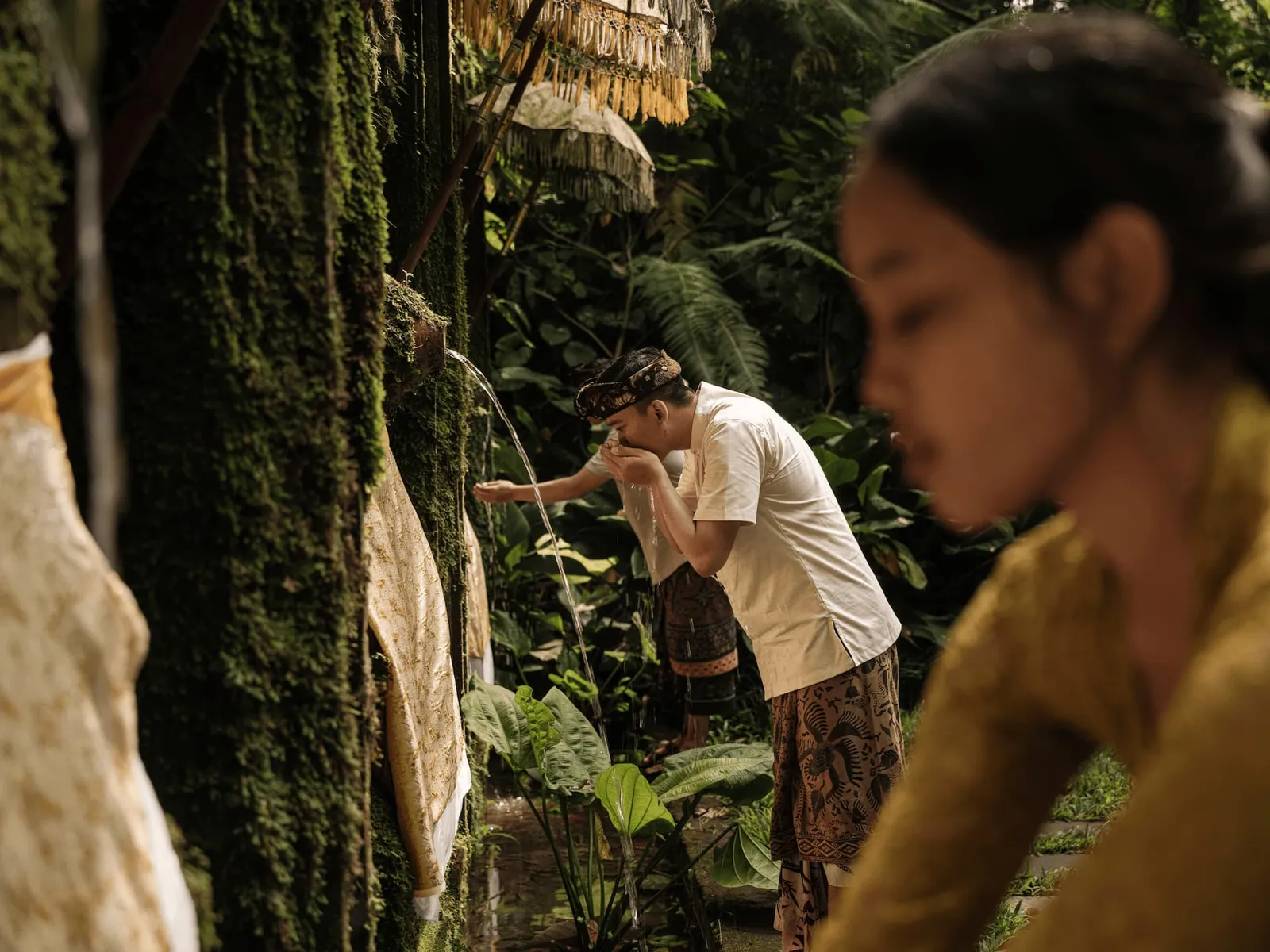 Melukat at the spring by the Wos River: a man in a sarong bathes his face under a spout of water, among moss and ceremonial parasols.