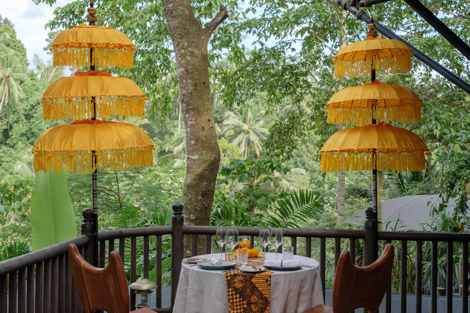 A table for 2 laid on a deck, between 2 tiered yellow ceremonial parasols and the valley’s trees.